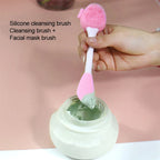 Silicone cleansing brush with a pink handle being used to scoop facial mask from a jar.