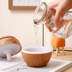 Hand pouring liquid from a glass container into a wooden cup on a light-colored surface.