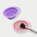 Purple and pink silicone cleaning dishes with a makeup brush on a white background